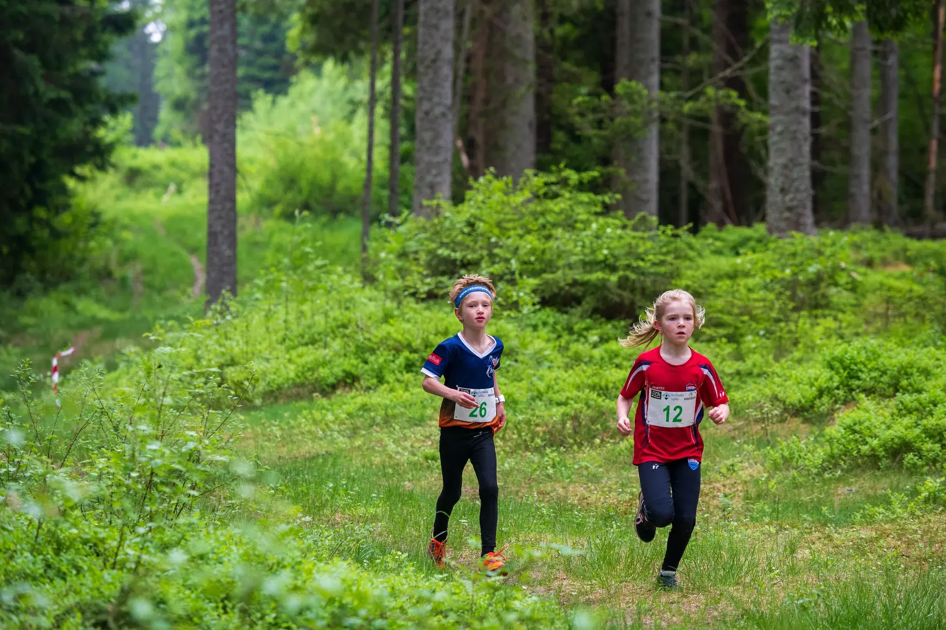 Två barn med nummerlappar springer sida vid sida under barnloppet, 2024