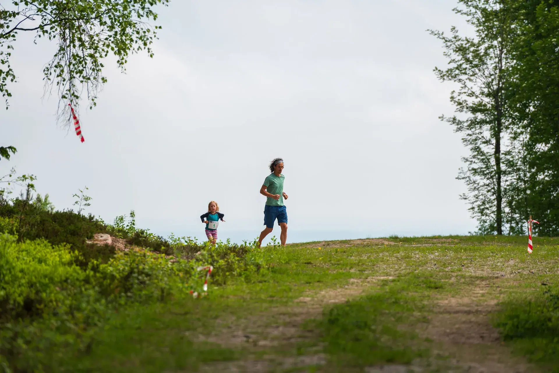 Ett barn och en vuxen kommer över krönet med himlen bakom sig under barnloppet, 2024