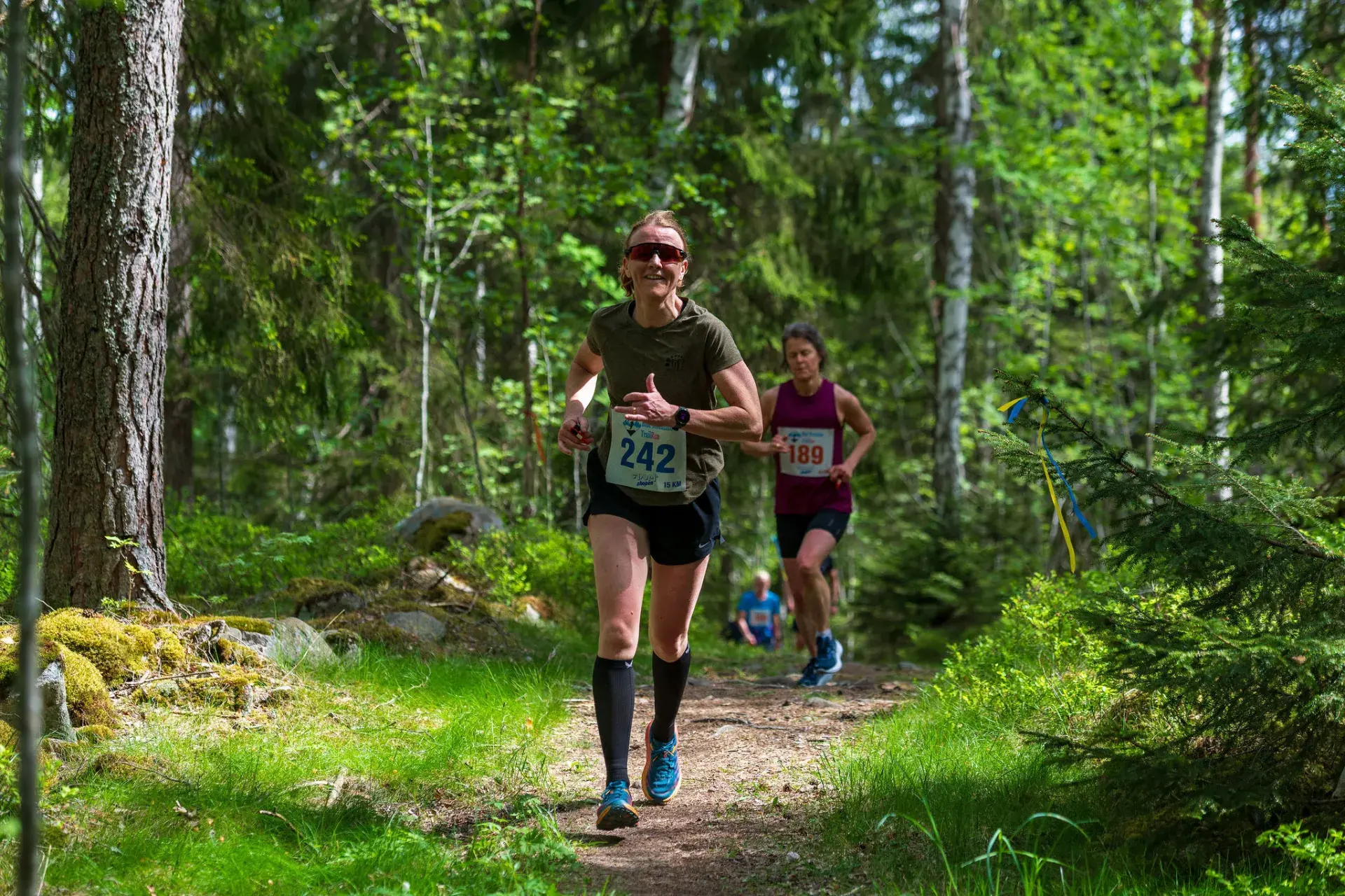 Leende löpare i solglasögon på stigen genom granskogen, med en löpare bakom sig, 2025